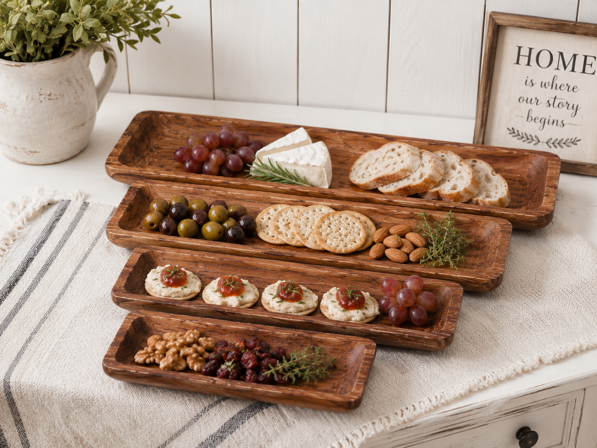 Four rustic rectangular wooden charcuterie boards arranged on a white tablecloth, styled with grapes, crackers, bread, nuts, cheese, and small appetizers in a bright kitchen setting.