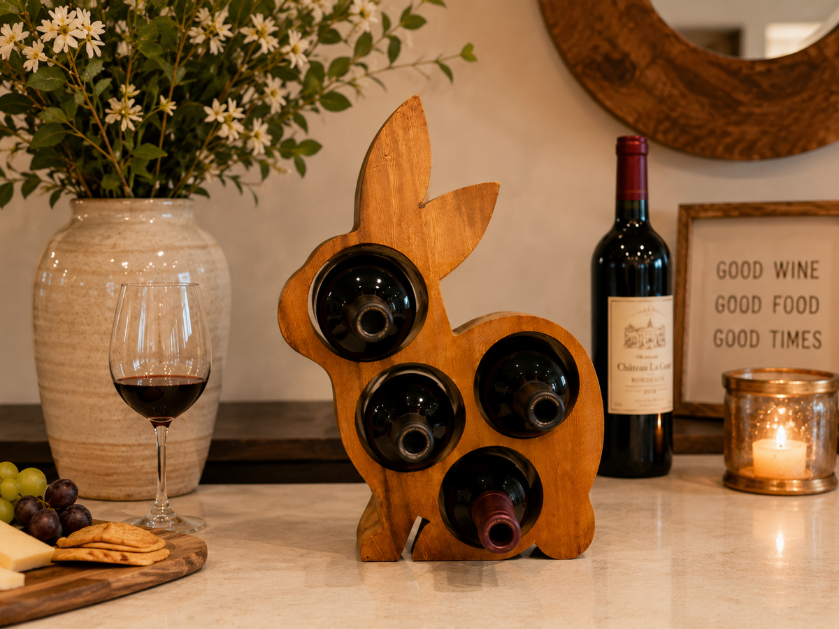 A wooden rabbit-shaped wine bottle holder on a kitchen counter, holding three wine bottles. A glass of red wine, a vase with white flowers, grapes, crackers, and a lit candle are arranged around it in a warm, home-style setting.