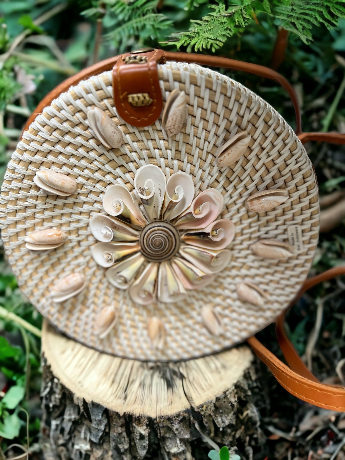 Round woven bag with sea shell floral design and brown strap on a natural background