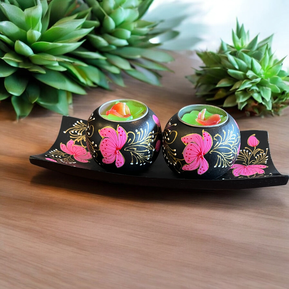 Two small black decorative candle holders with pink butterfly and gold floral designs, each containing a green tea light candle, displayed on a black tray on a wooden surface with green plants in the background.