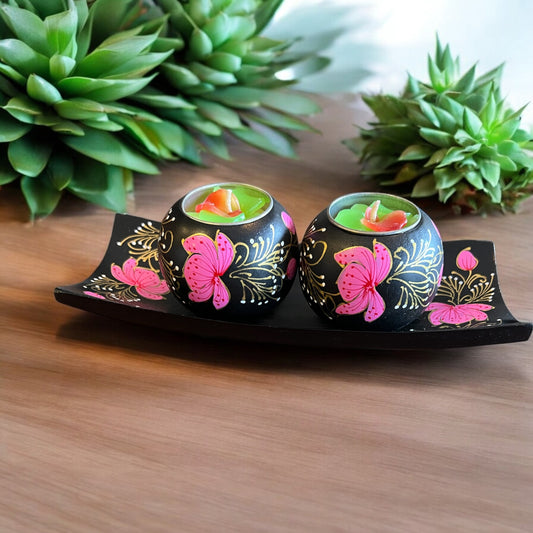 Two small black decorative candle holders with pink butterfly and gold floral designs, each containing a green tea light candle, displayed on a black tray on a wooden surface with green plants in the background.