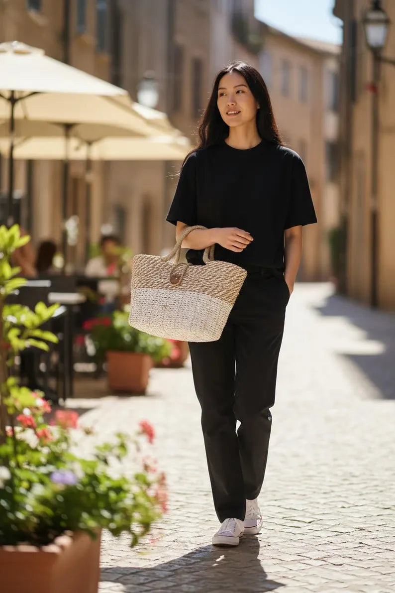 Woman walking down a sunlit street holding a woven bag