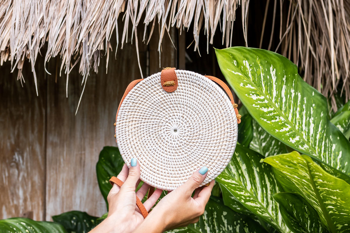 Round woven bag held by a hand with green leaves and straw roof in the background