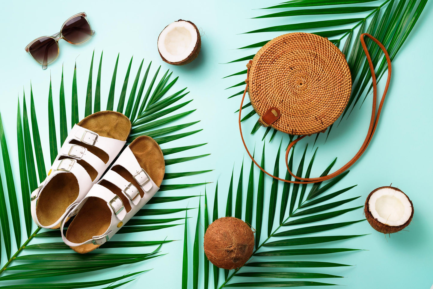 Summer accessories including sandals, a hand woven round rattan bag, coconuts, and sunglasses on a light blue background with palm leaves.